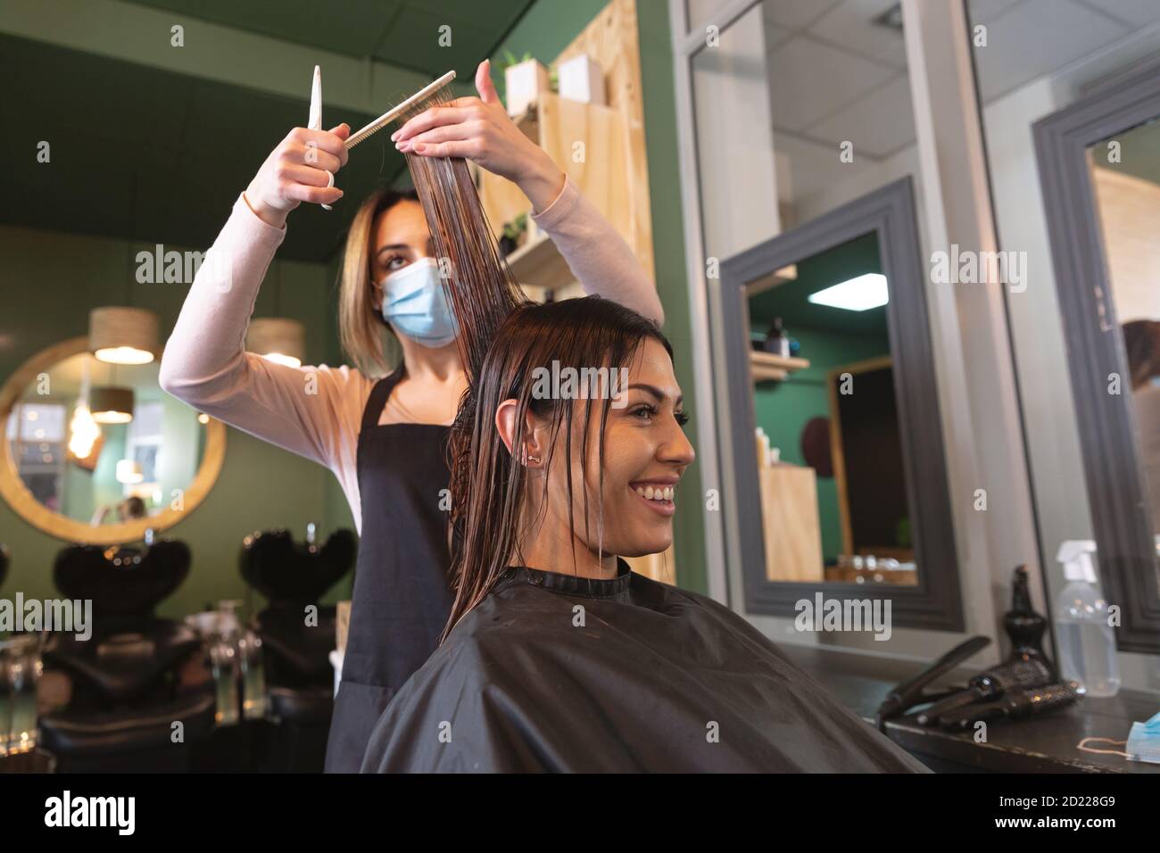 Female hairdresser wearing face mask cutting hair of female customer at hair salon Stock Photo