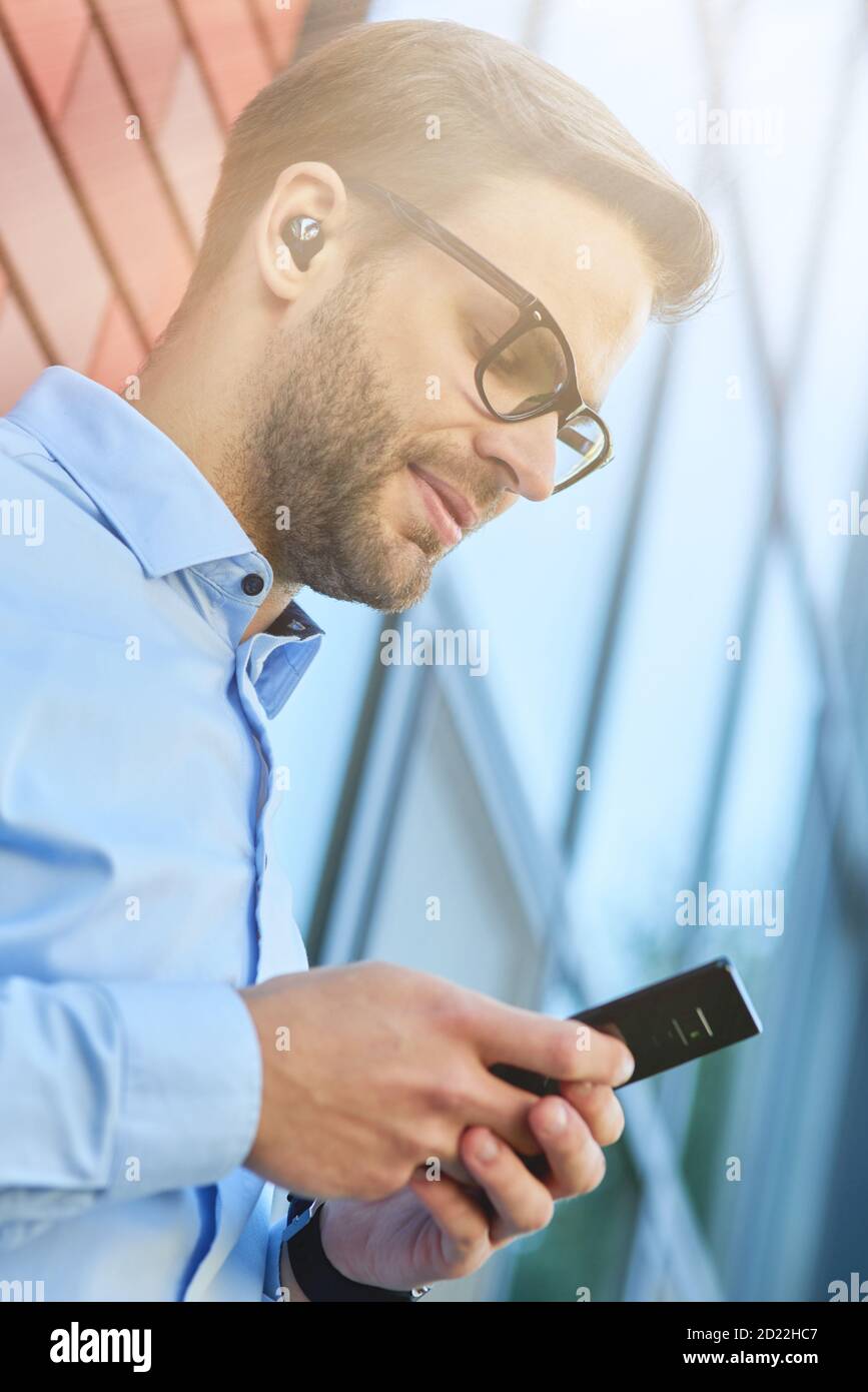 Side view of a young handsome businessman wearing wireless earphones using his smartphone while leaning against brick wall outdoors Stock Photo