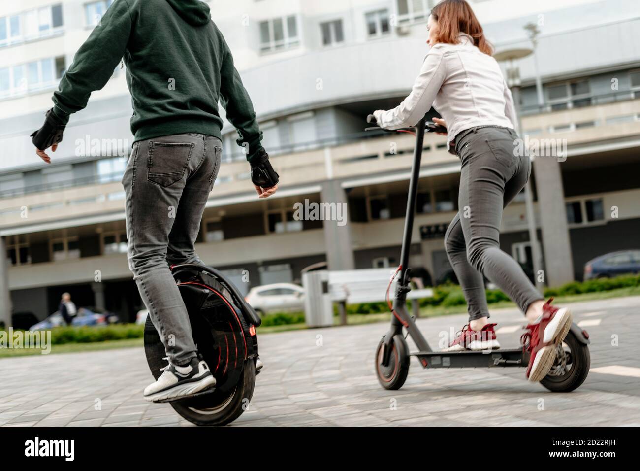 Young man riding unicycle and young woman in casual wear riding on electric kick scooter on city street Stock Photo