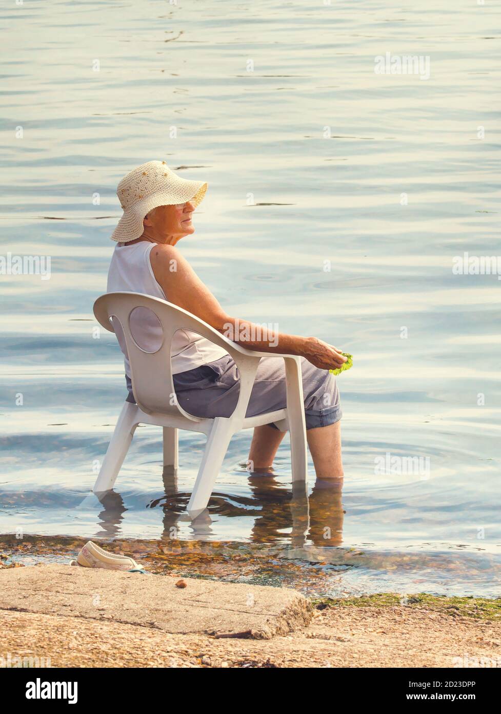 Elderly Lady In A Straw Hat With Shoes And Socks Off And Trousers Rolled Up Sitting On A Plastic Chair In The Sea Cooling Off In The Summer Sun. Stock Photo