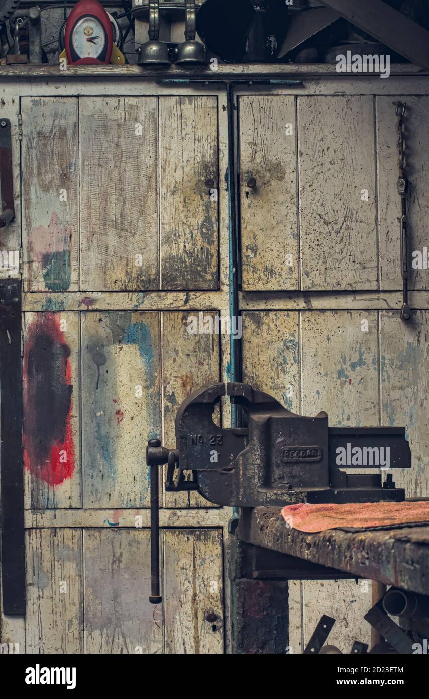 Old Ancient Record Vice In Front Of A White Distressed Dirty Paint Spattered Old Cupboard In A Cluttered Old Workshop. Mudeford UK Stock Photo