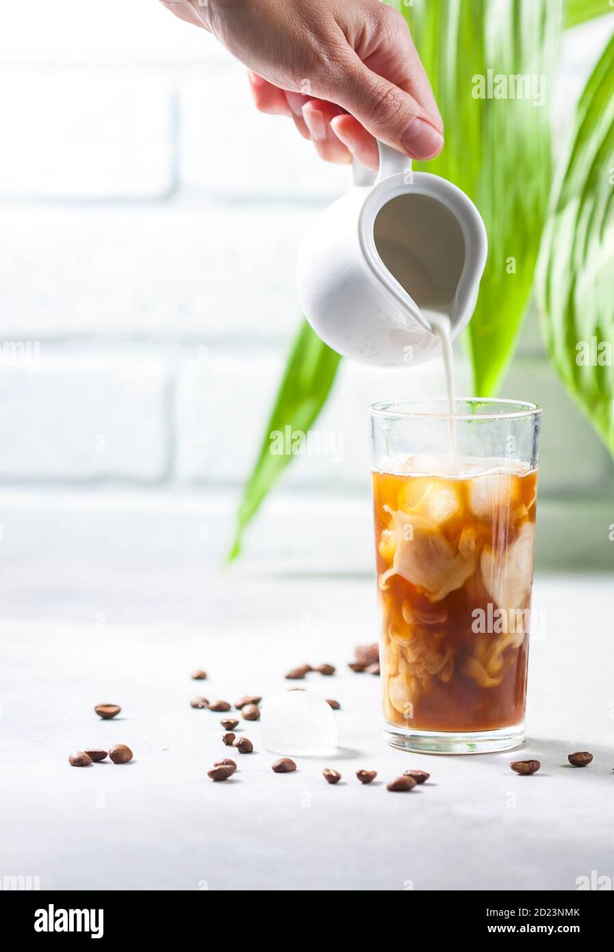 Cold brewed iced coffee in a tall glass cup and coffee beans on a gray concrete background. Pouring milk into a glass of coffee. Stock Photo