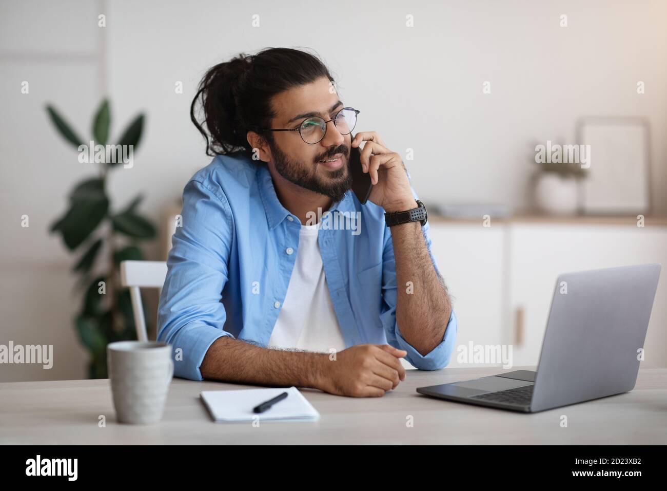 Millennial Indian Man Talking On Cellphone And Using Laptop At Home Office Stock Photo