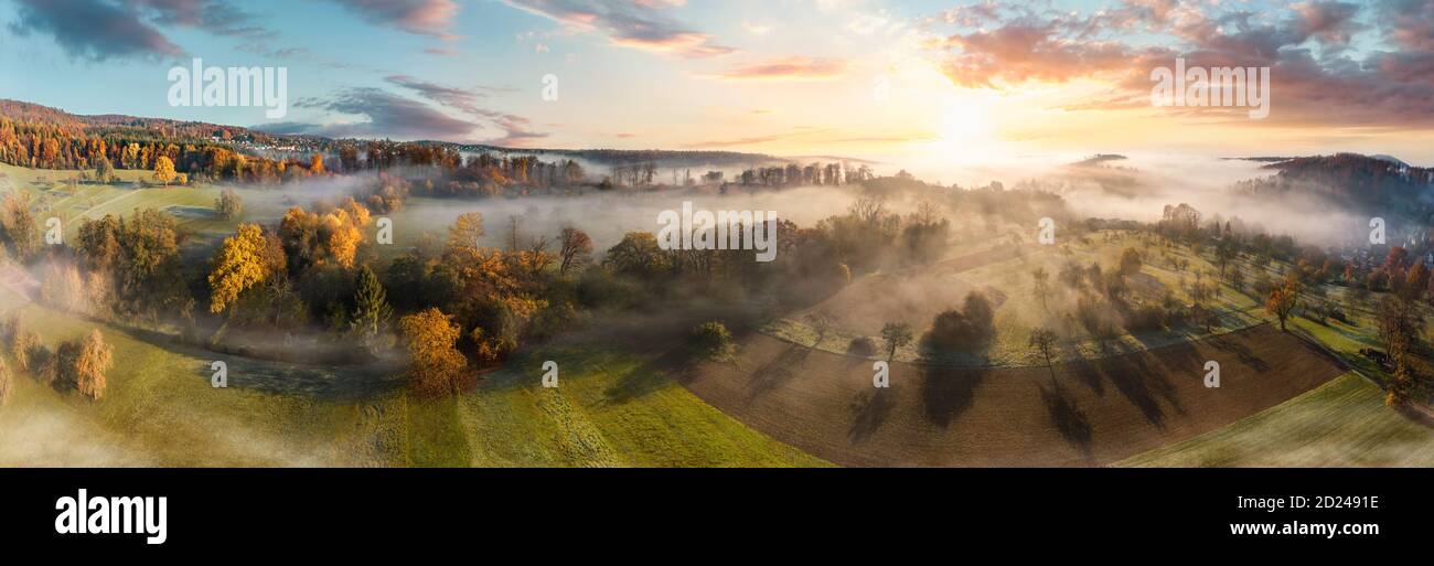 Stunning aerial panorama of a misty rural landscape at sunrise, with colorful autumn trees, fields and meadows, blue sky, trees casting shadows and be Stock Photo