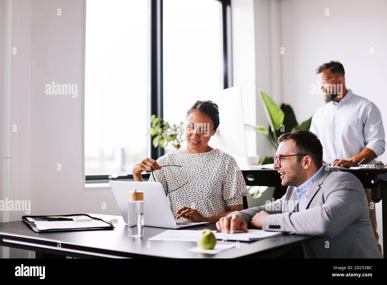 Two smiling businesspeople discussing work on a laptop at an office desk with a colleague working in the background Stock Photo