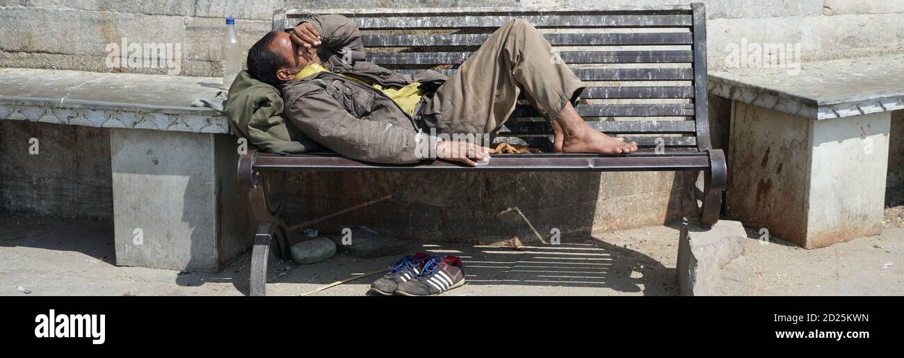 Poor homeless beggar man or refugee sleeping on a dirty wooden bench in a one-way street in the city during day time. Social documentary concept. Shoe Stock Photo