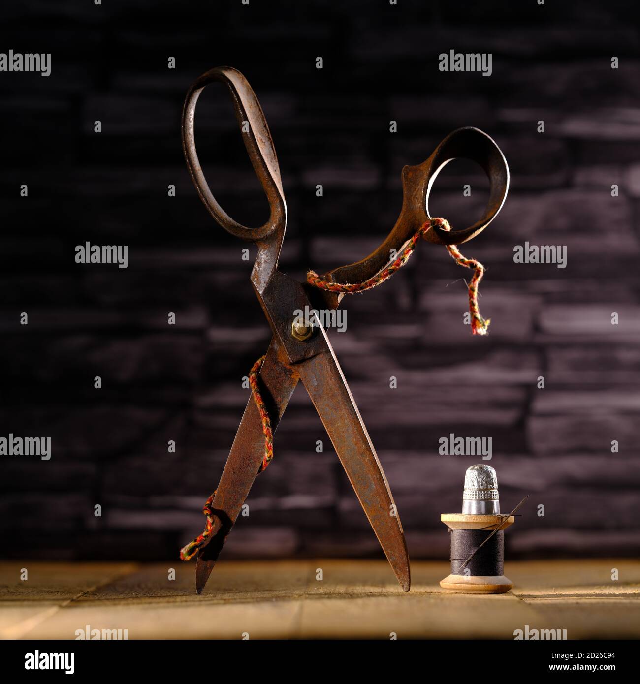 Rusty vintage pair of scissors balanced on timber flooring with black cotton reel thread needle and silver thimble against slate background Stock Photo