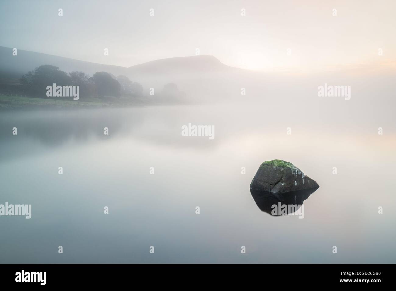 Embsay Crag peeks through the fog floating over Embsay Reservoir on a calm September morning with low visibility creating a minimal and tranquil scene. Stock Photo