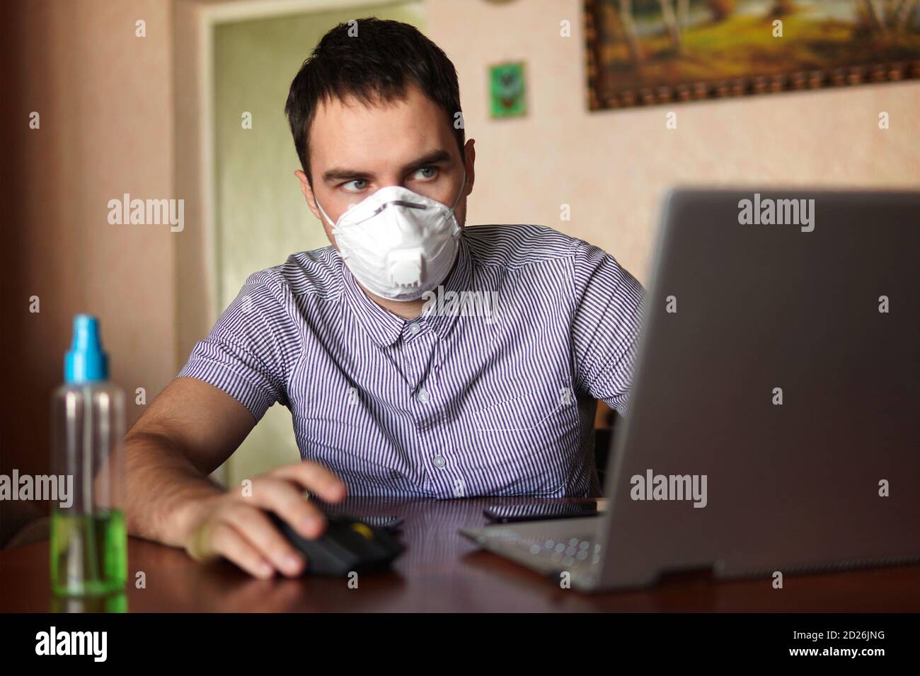 young man wearing medical face mask at work from home office due to corona virus outbreak with laptop on wooden table, remote work against background Stock Photo