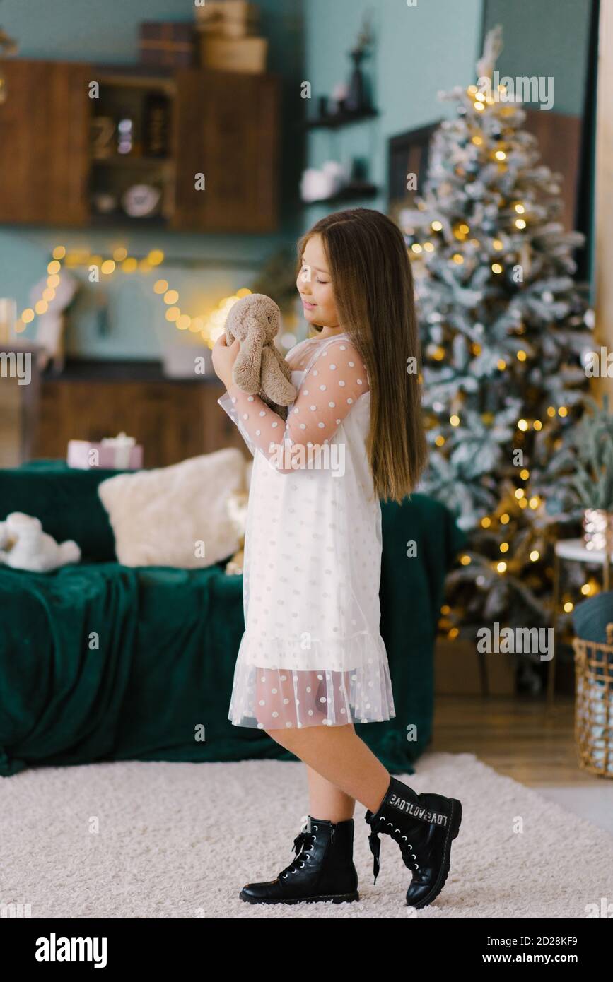 The concept of a happy New year. A baby girl near the Christmas tree holds a Teddy bear in her hands against the background of the Christmas tree. Bes Stock Photo