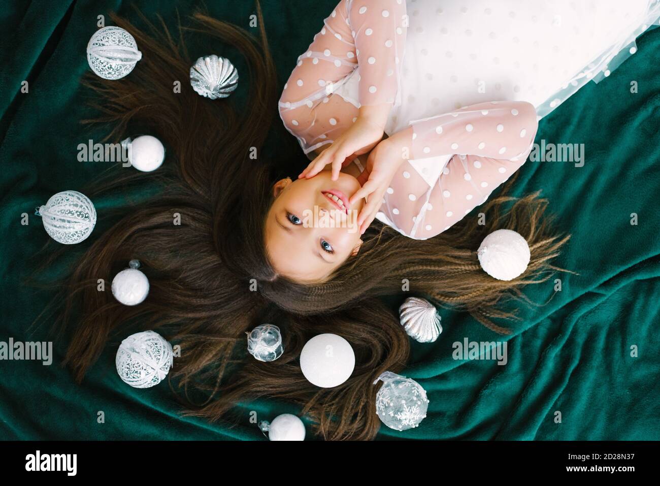 A beautiful girl with long hair is lying on her back on a green blanket. On her hair are white Christmas balls. Happy Christmas and New Year concept Stock Photo