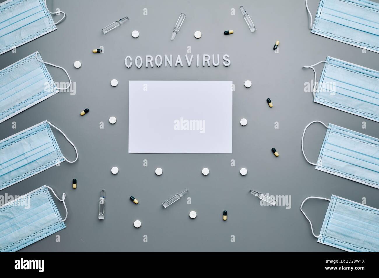 Above view composition of medical masks framing blank white paper with coronavirus word and medication laid out over grey background, copy space Stock Photo