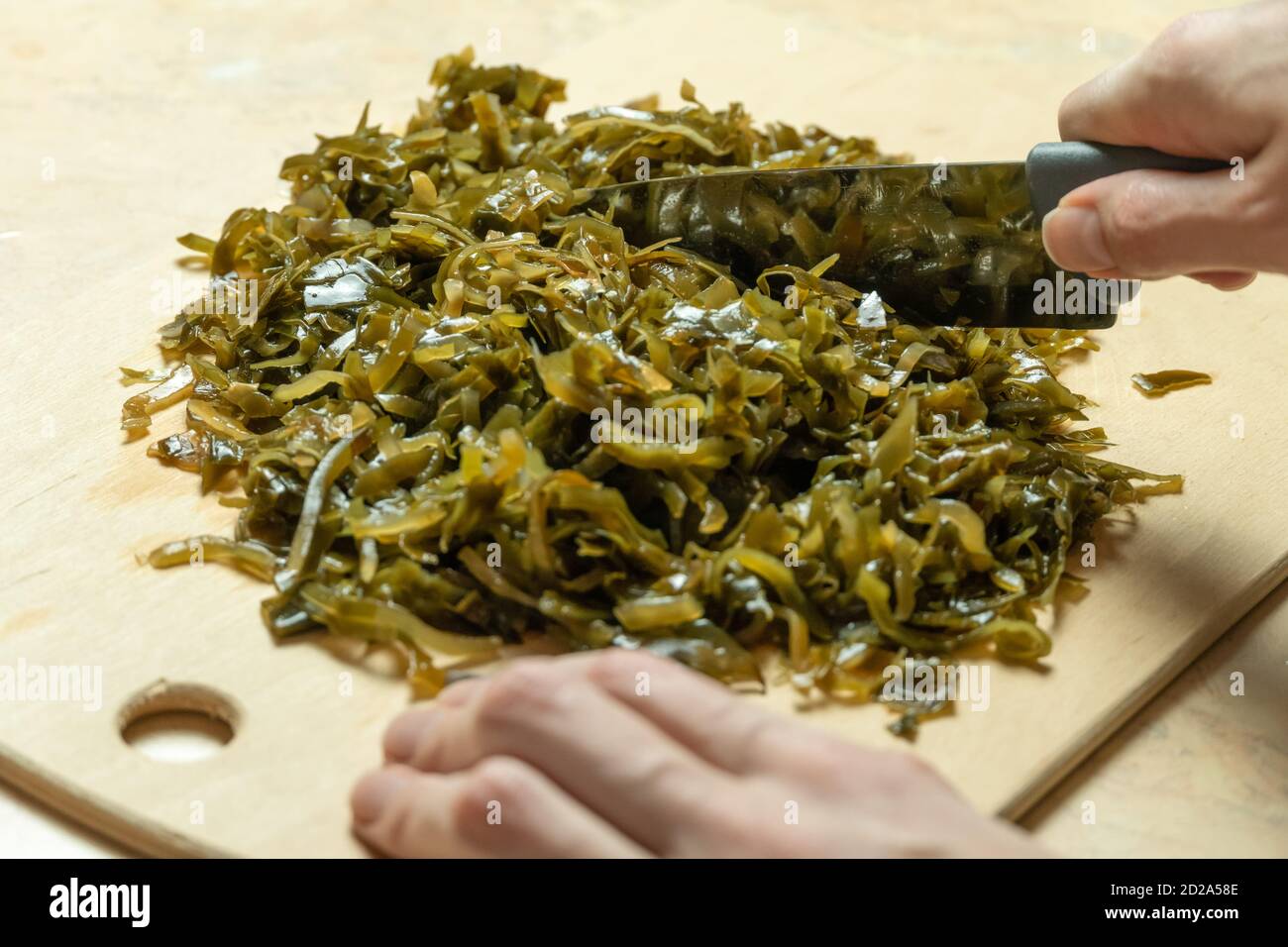 Women's hands cut green seaweed Laminaria on a wooden Board with a kitchen knife close up Stock Photo