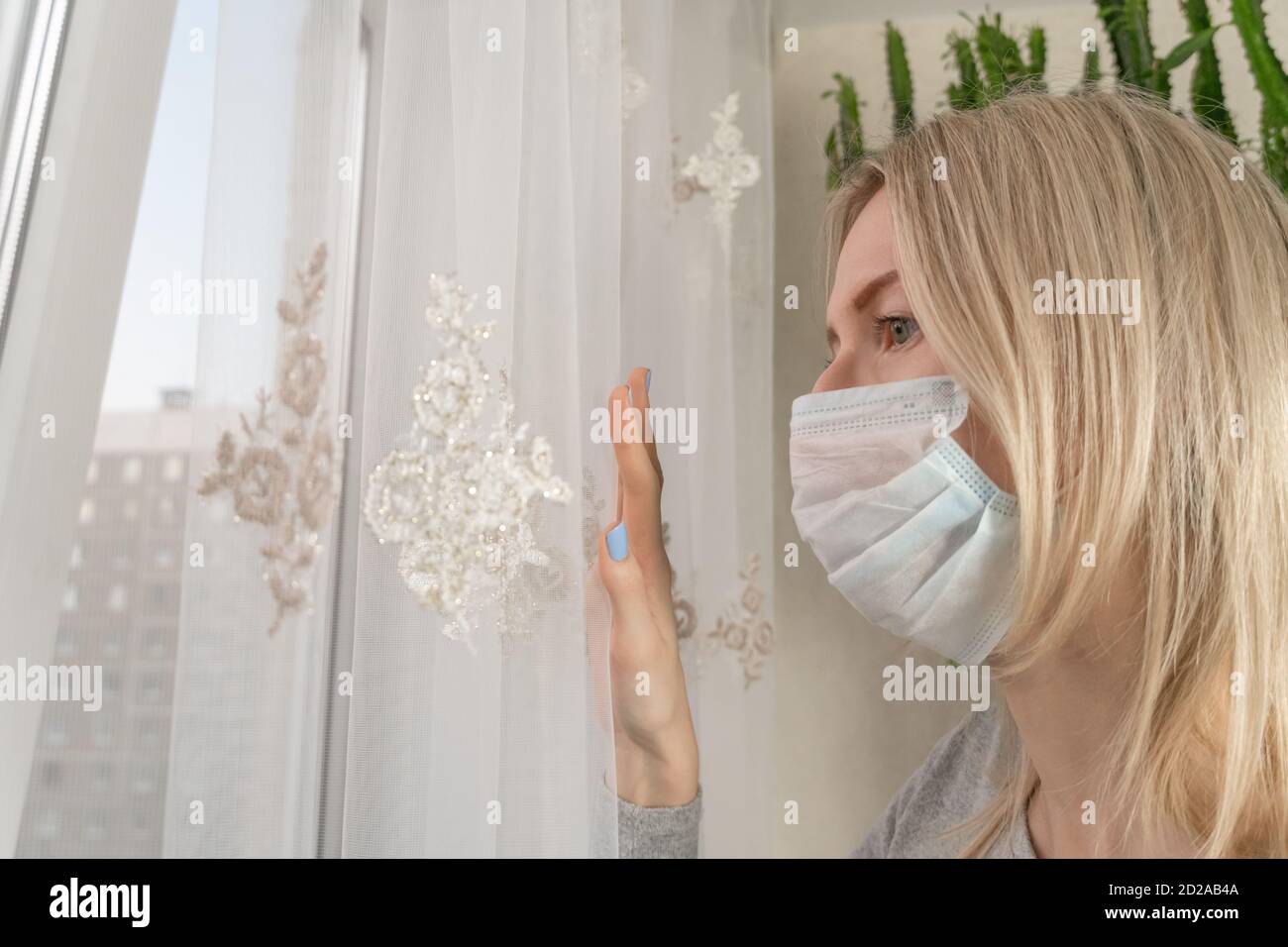 Sad blonde girl in a medical mask isolated at home looking out the ...