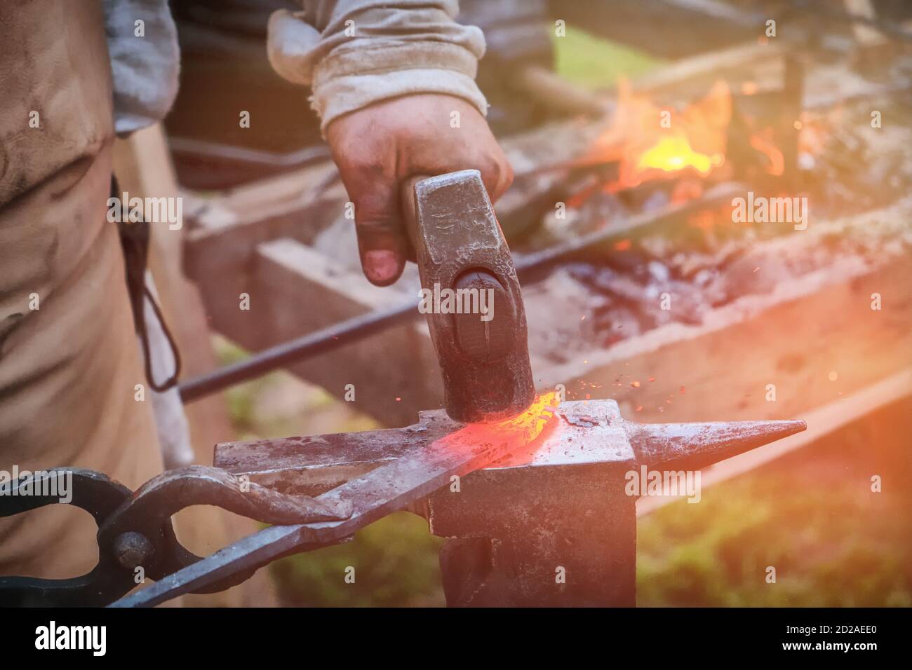 The blacksmith strikes the red blade of the sword with a hammer. Work in the forge to create weapons. The knife is between a rock and a hard place. Stock Photo