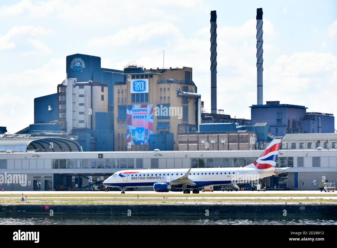 British Airways jet plane taxiing to take off at London City Airport Tate & Lyle Silvertown sugar refinery factory buildings beyond Newham England UK Stock Photo