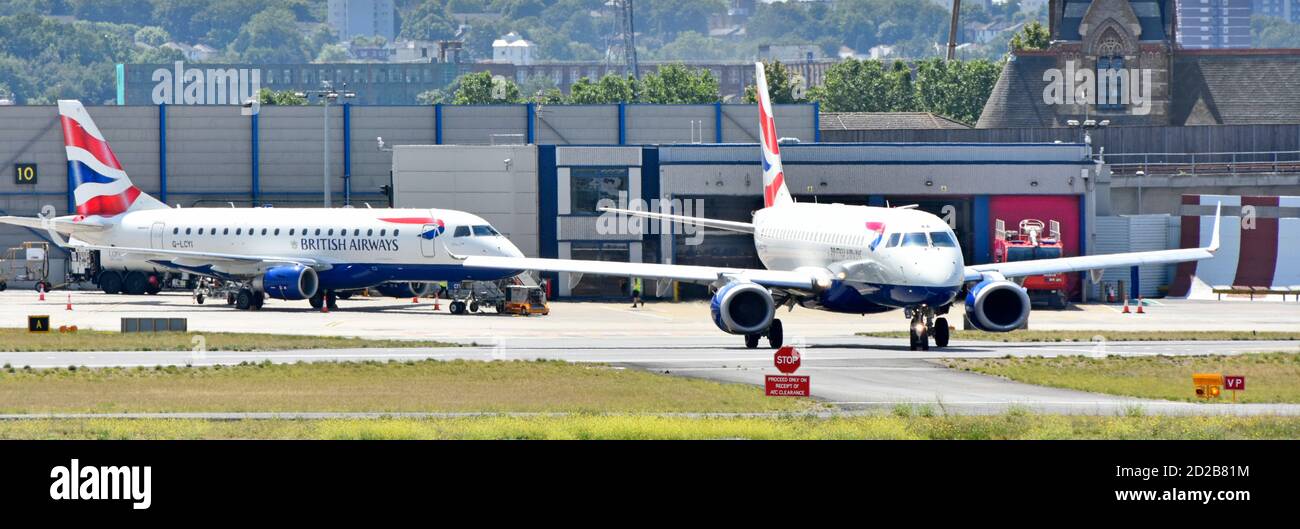 British Airways jet plane taxiing to take off East London City Airport with fire engine in fire station building beyond Silvertown London England UK Stock Photo