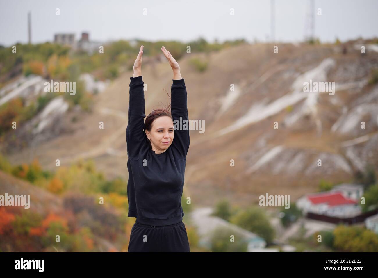 Young woman doing warm-up in hilly terrain. Adult female in black casual clothes doing sports in fresh air in countryside. Stock Photo