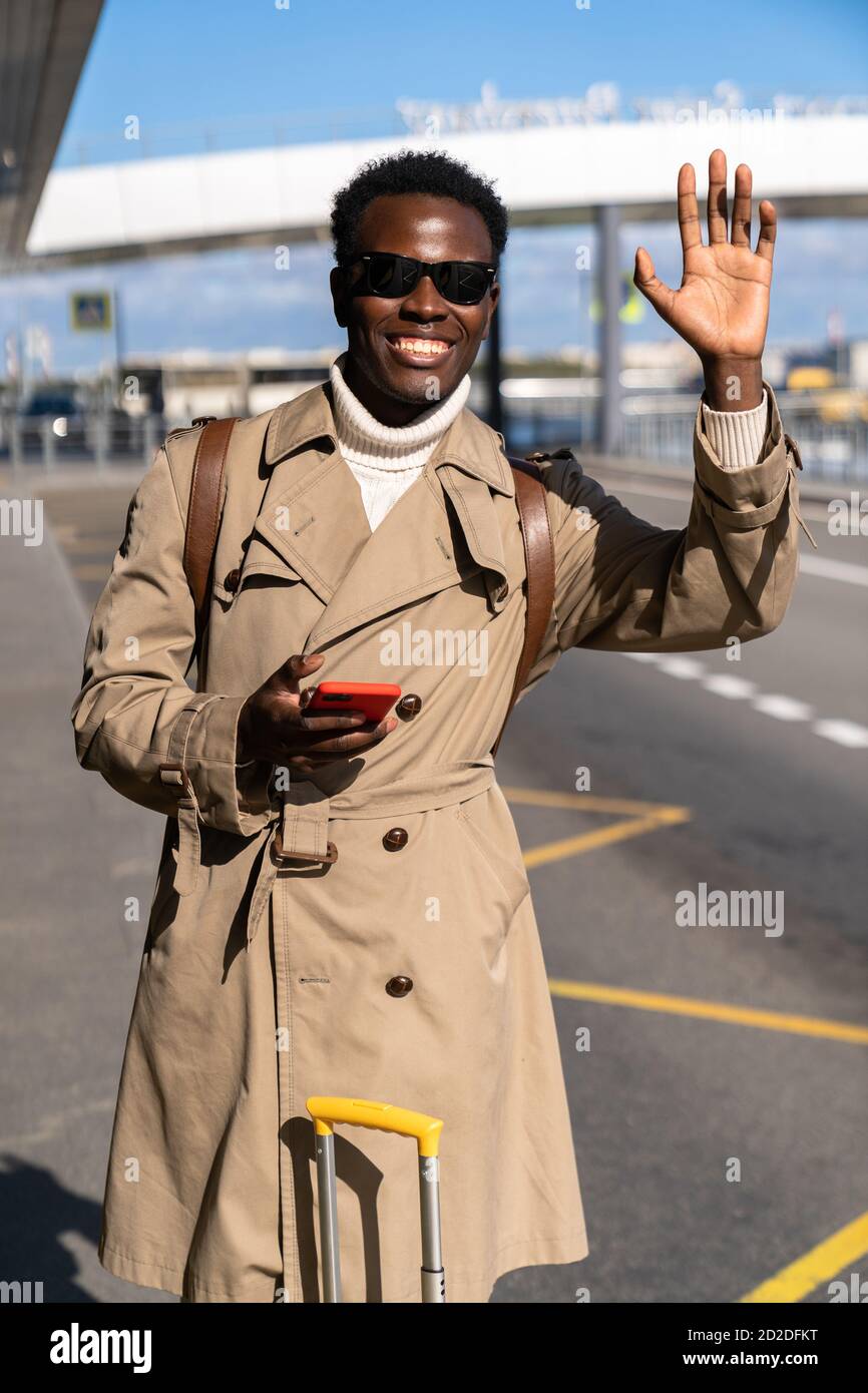 Afro-American millennial traveler man wear beige trench coat with suitcase stands in airport terminal, holding mobile phone, calling taxi, raising han Stock Photo