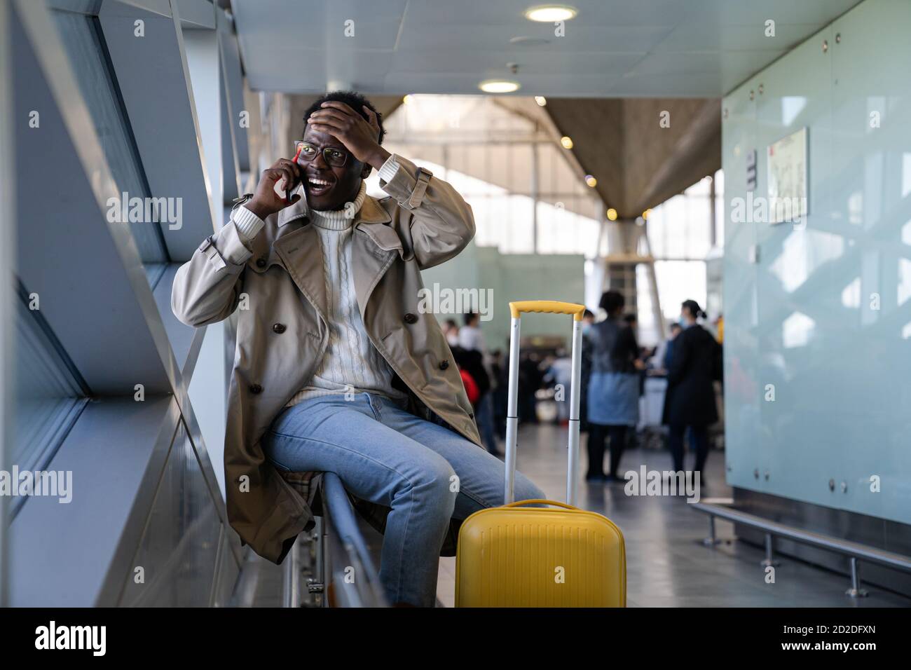 Stressed Black man in spectacles shocked with bad news, talking on mobile phone, sitting in airport, makes facepalm by hand and forgot something impor Stock Photo