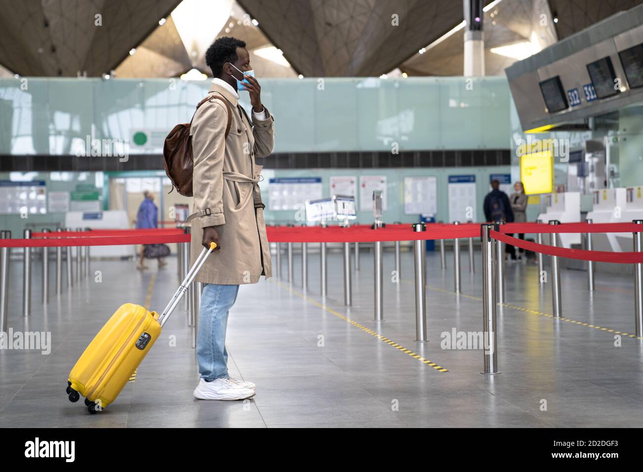 Black man with suitcase wearing face protective mask during virus epidemic, covid-19 pandemic while standing at almost empty airport terminal. Second Stock Photo