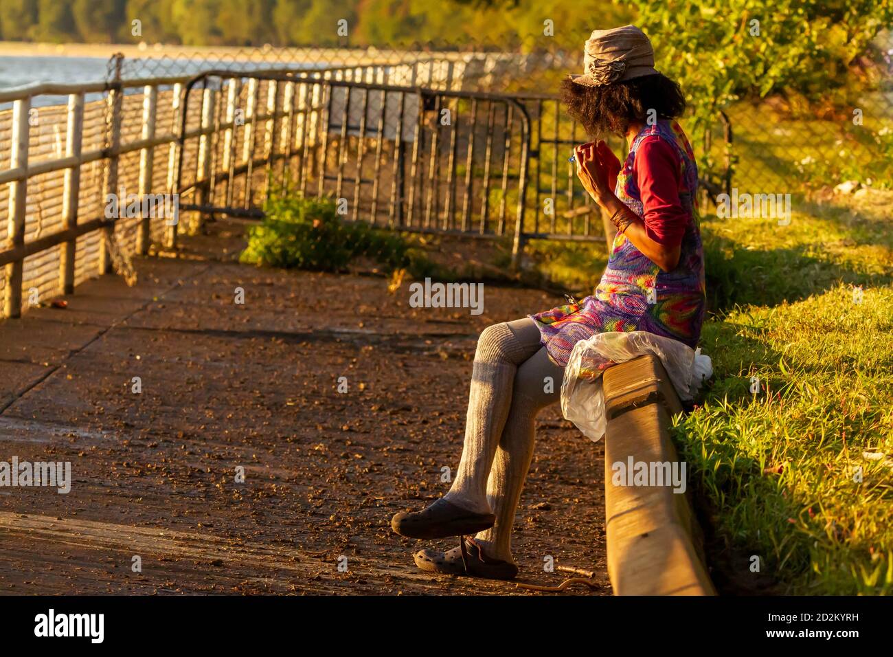 An African American woman wearing long gray socks and vibrant colored cotton dress is sitting crosslegged on a barrier wall by side walk playing with Stock Photo