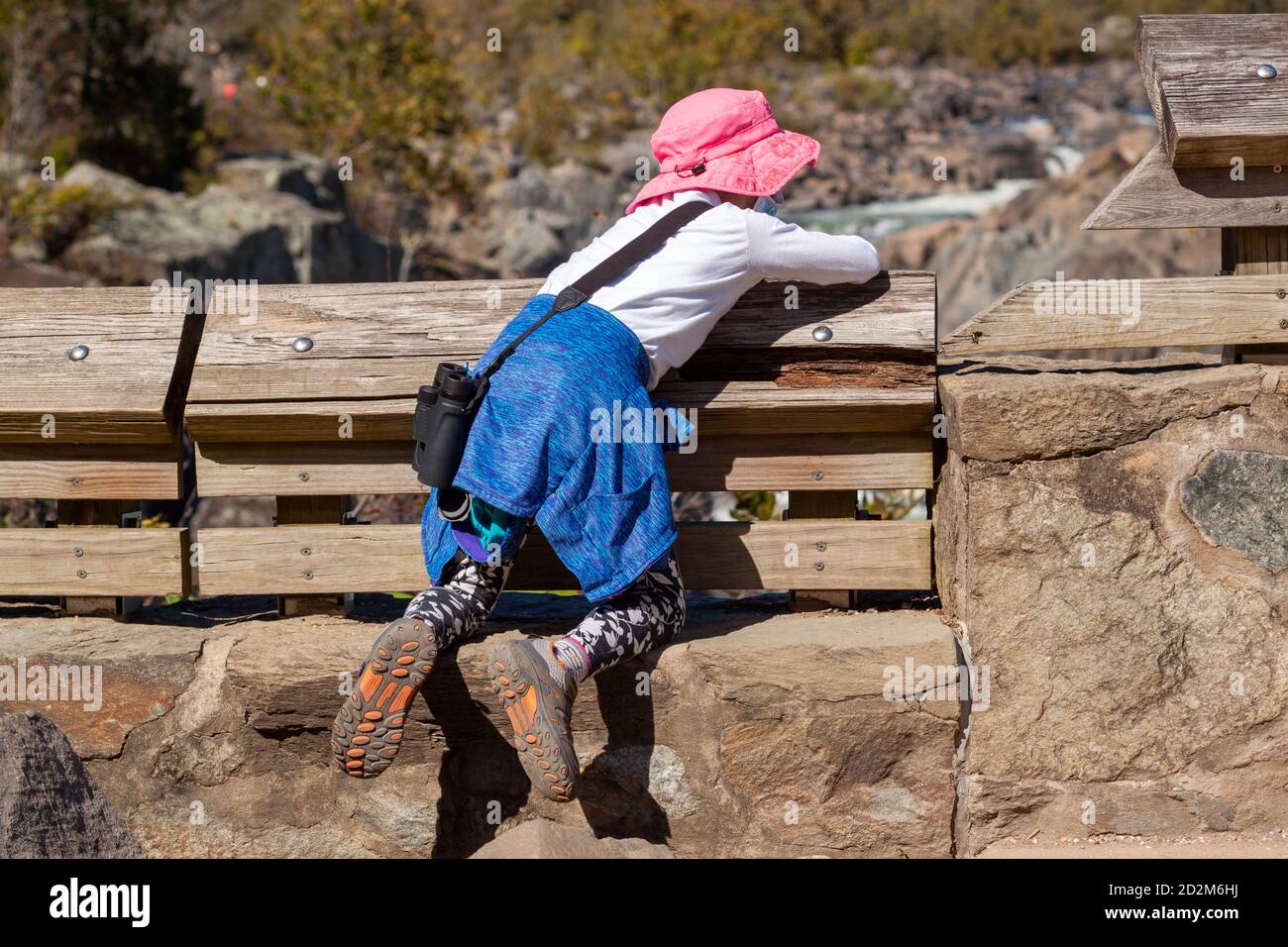 A curious little girl wearing pink solar hat and sneakers is climbing on the stone barriers to see the great falls of Virginia. This is a dangerous ac Stock Photo