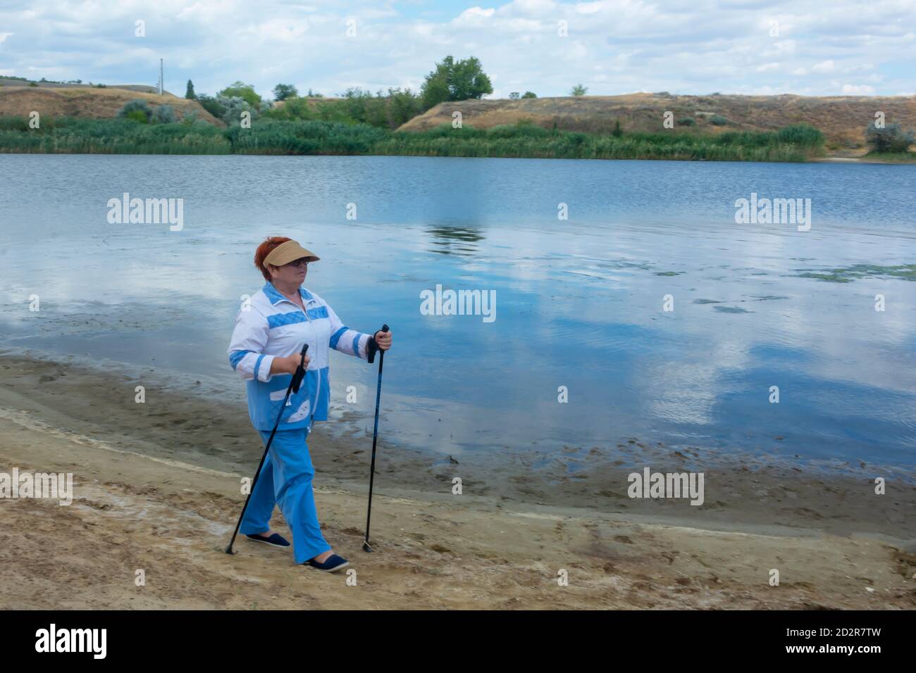 Scandinavian walking, an elderly woman walks along the river bank with sticks. Active lifestyle concept in old age. Soft focus, blur. Stock Photo