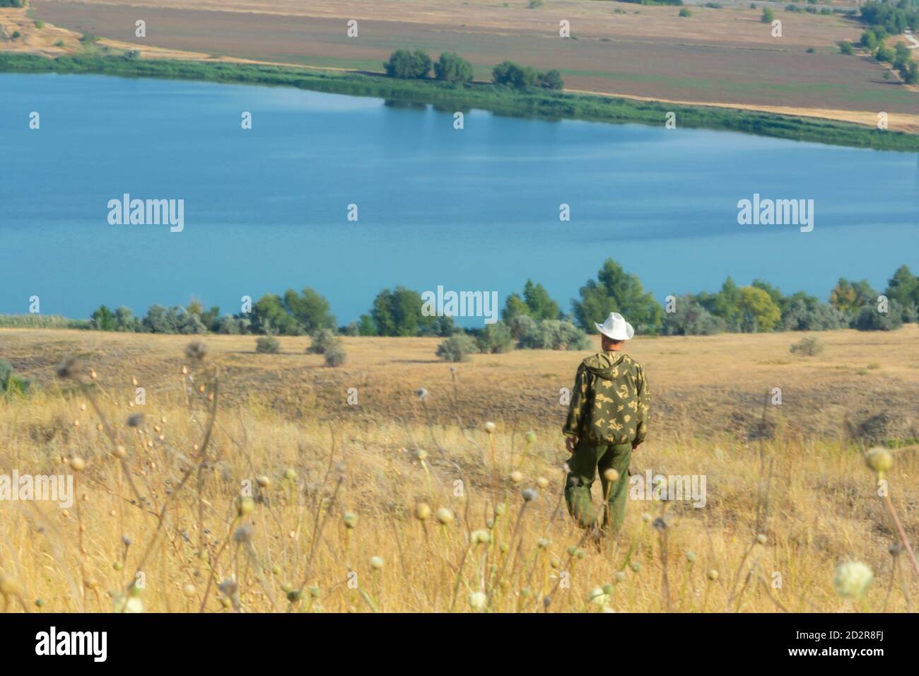 Man in hat goes to river in summer. Beautiful landscape of nature. Volga in the steppe, feather grass and grasses on shore. Soft focus, fog view. Stock Photo