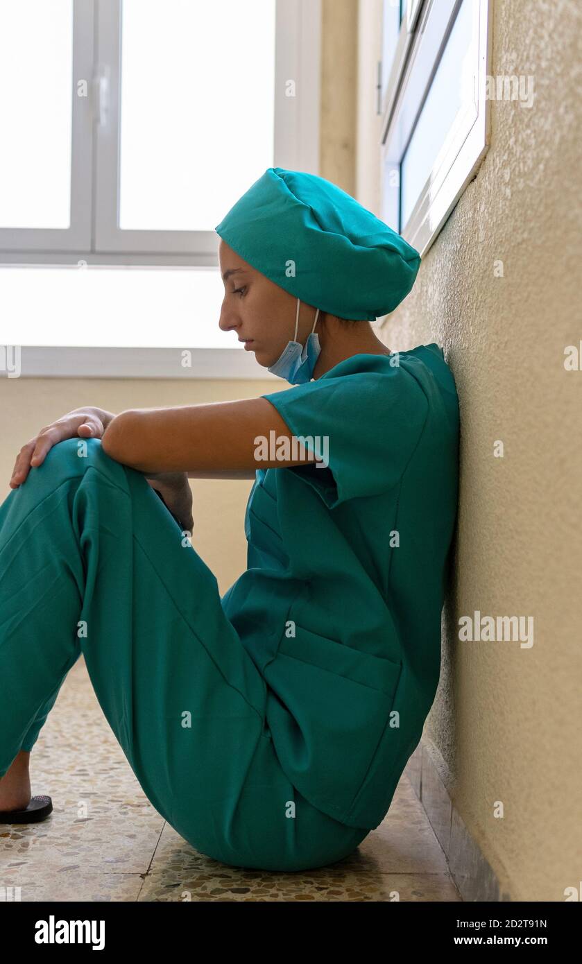 Side view of exhausted young female medical worker sitting on floor near wall after difficult work in hospital during coronavirus pandemic Stock Photo