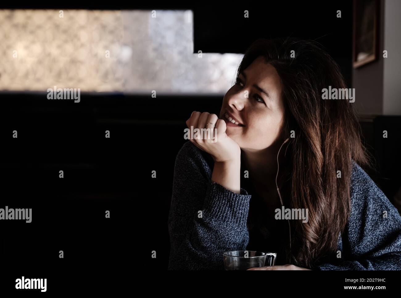Dreamy female sitting at table in cafe and listening to music in earphones while leaning on hand and thoughtfully looking away Stock Photo