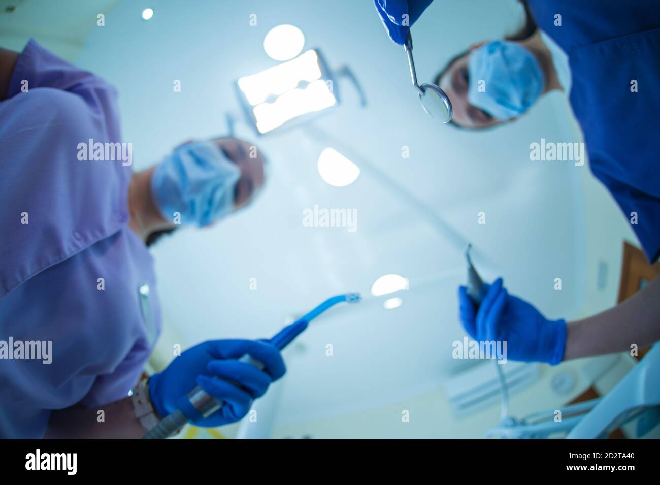 Blurred female dentist with mirror and drill and assistant with suction device standing against