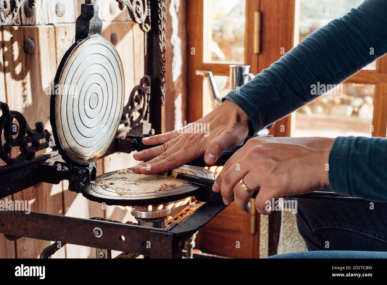Cropped unrecognizable artisan using stick while rolling up hot freshly baked Catalan neula waffles on waffle iron placed over gas flame Stock Photo