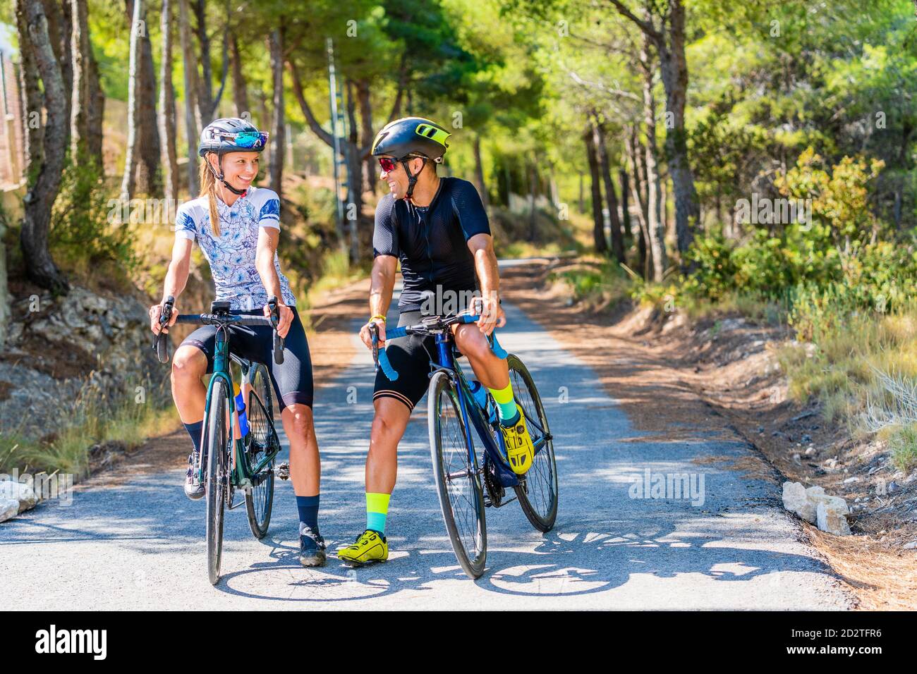 Full body of happy sportive man and Woman in activewear and helmets standing with bicycles on asphalt roadway and resting during ride through mountain Stock Photo