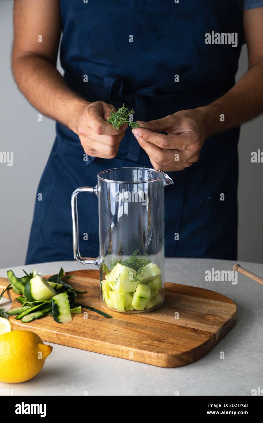 Crop unrecognizable male adding fresh green herb into blender cup with chopped cucumber while preparing healthy smoothie in kitchen Stock Photo
