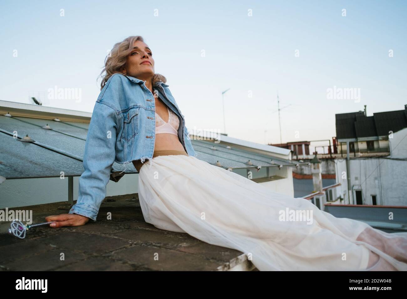 Tranquil female in denim jacket and with wavy hair relaxing in house roof in the city in evening and looking away Stock Photo