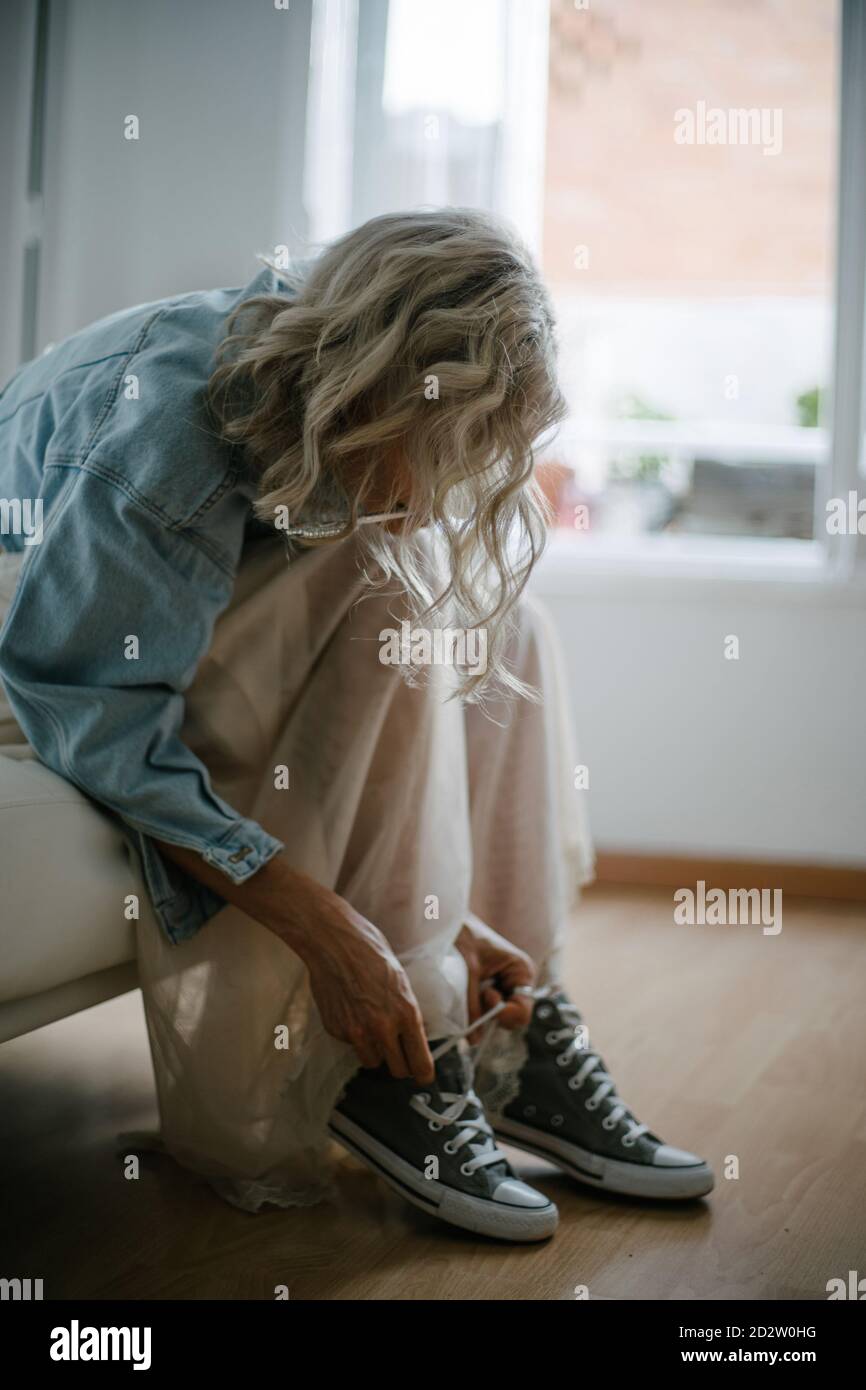Side view of stylish female in denim jacket and skirt sitting on sofa and tying shoelaces on gumshoes Stock Photo