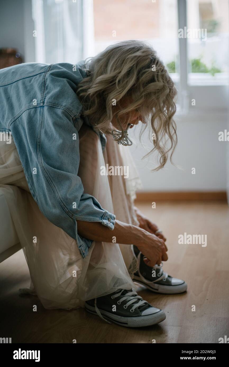 Side view of stylish female in denim jacket and skirt sitting on sofa and tying shoelaces on gumshoes Stock Photo