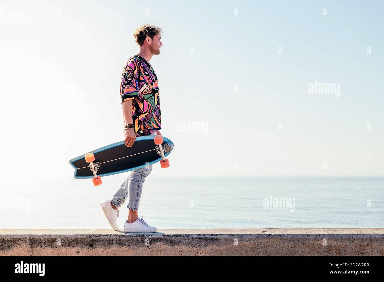 Low angle side view of young hipster male skater in trendy summer outfit  carrying skateboard while walking on embankment against sea and cloudless  sky in summer day Stock Photo - Alamy