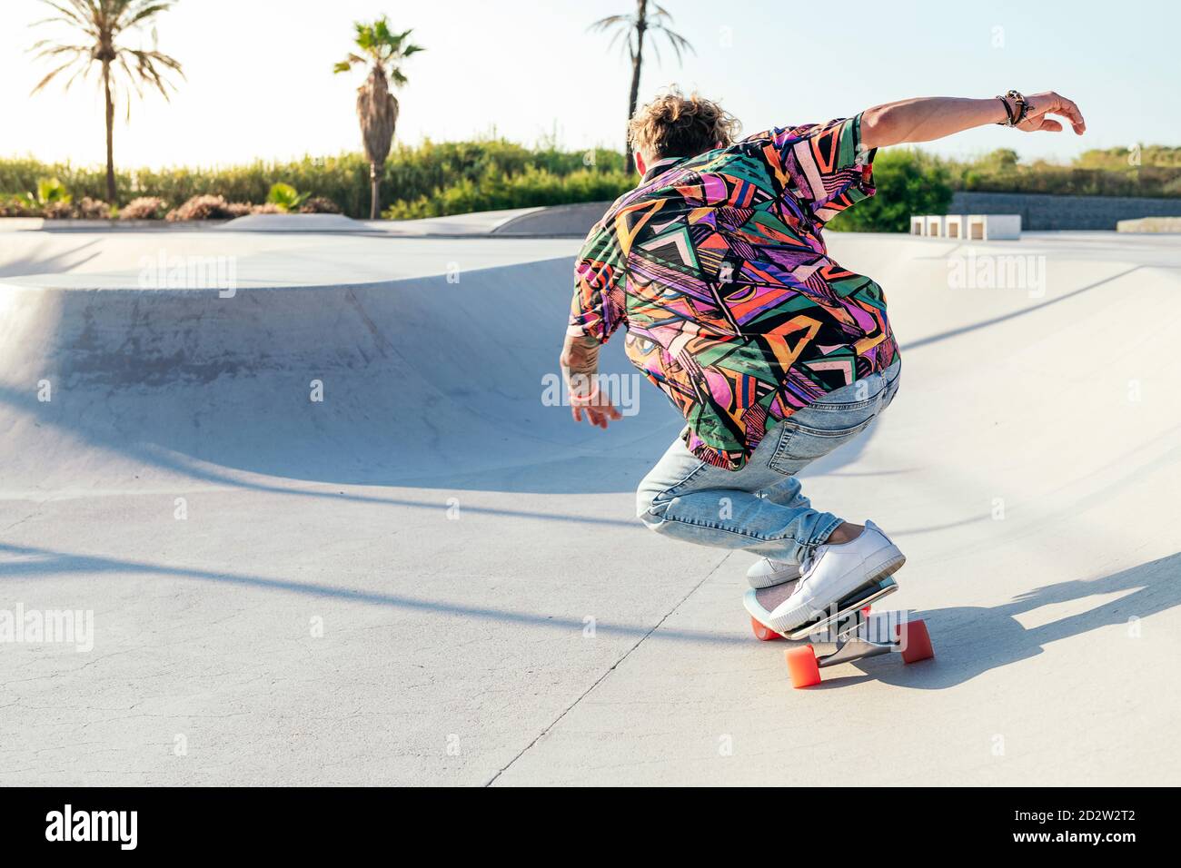 Back view of anonymous funky young male skateboarder in trendy colorful shirt and jeans performing trick on concrete ramp while practicing skills in skatepark Stock Photo