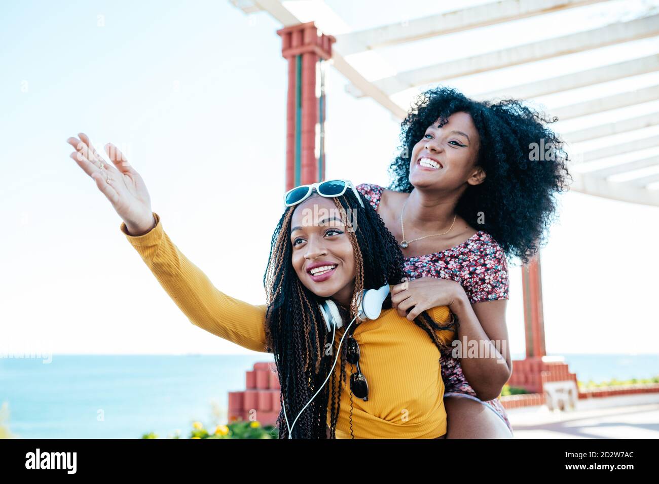 Cheerful ethnic Woman with curly hair piggybacking delighted female friend with braids while having fun on promenade at weekend in summer Stock Photo