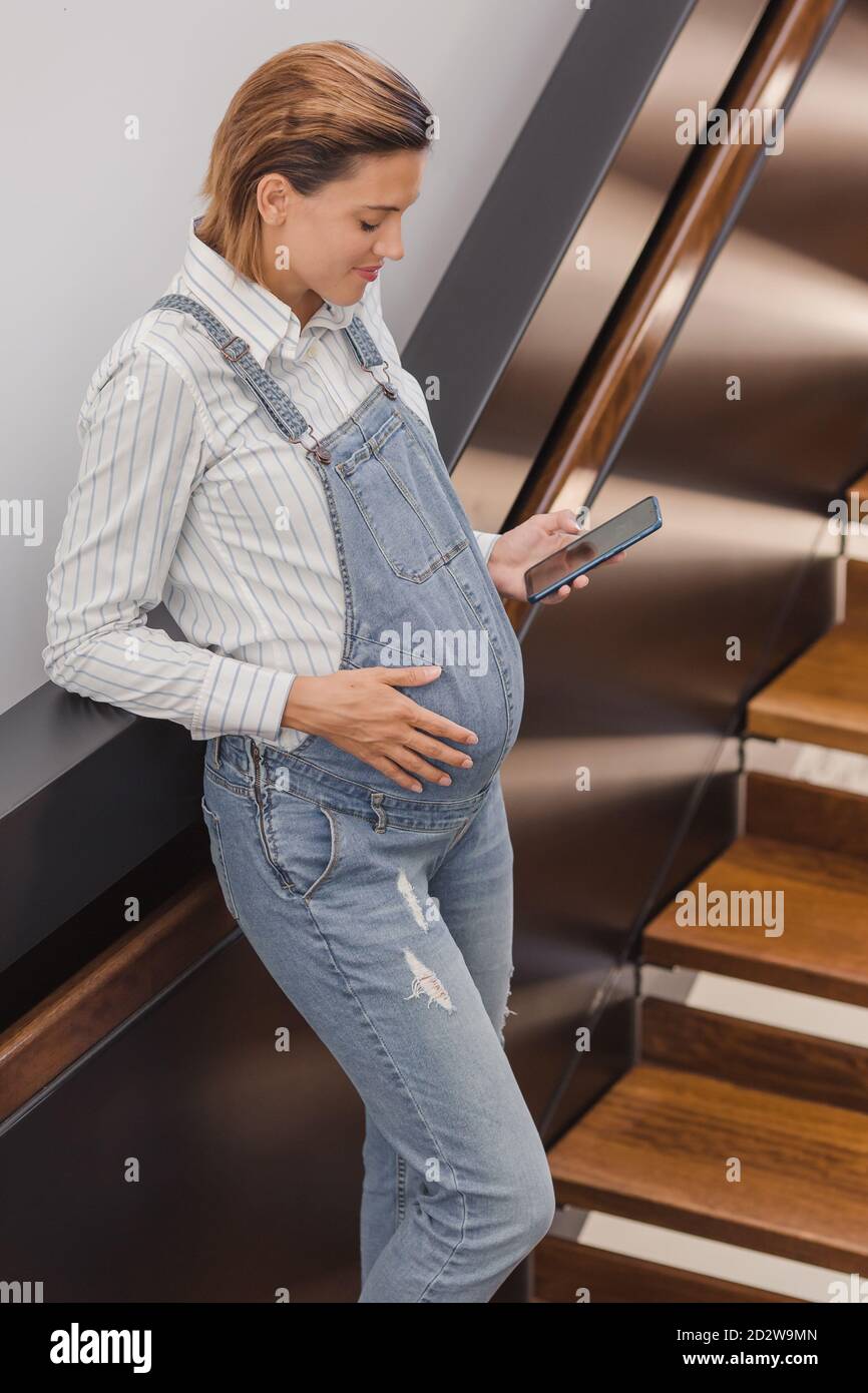 Concentrated pregnant female in denim overalls standing near staircase