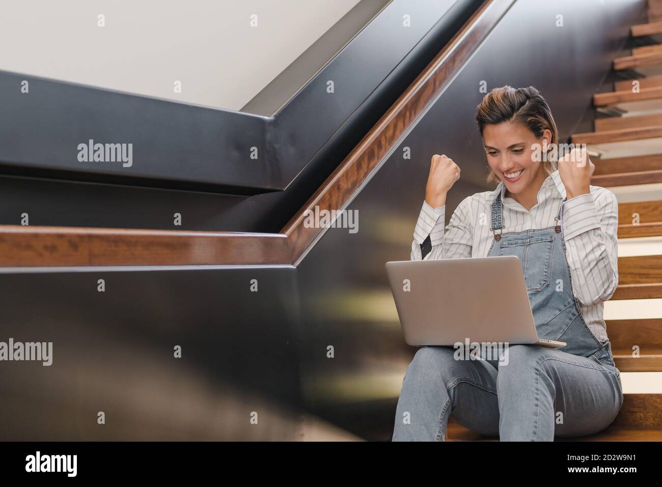 Delighted female entrepreneur celebrating goal achievement with fists up while sitting at netbook and working on project Stock Photo