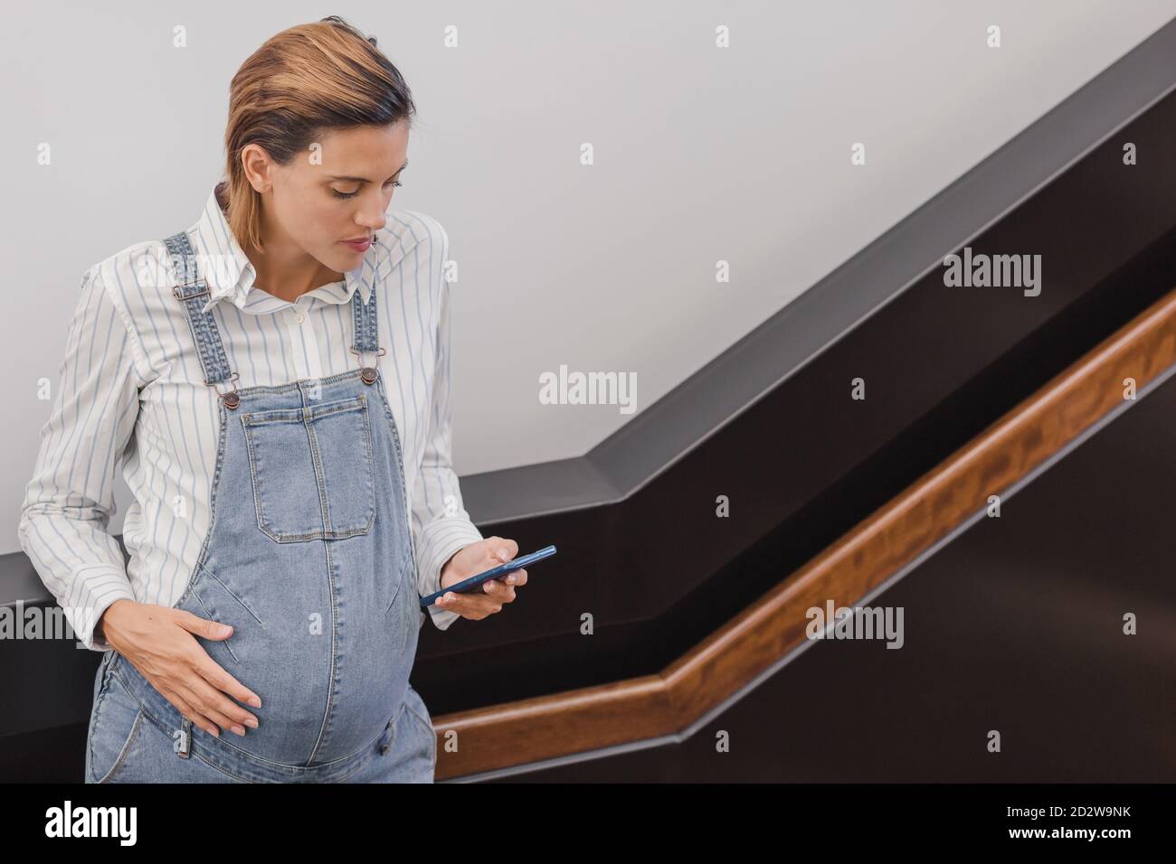 Concentrated pregnant female in denim overalls standing near staircase