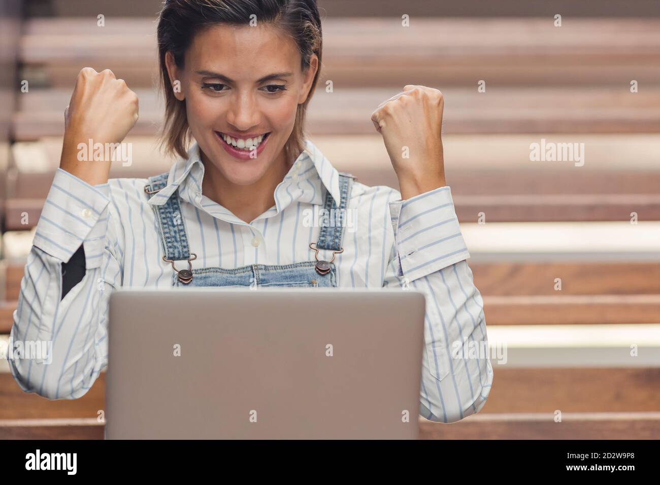 Delighted female entrepreneur celebrating goal achievement with fists up while sitting at netbook and working on project Stock Photo