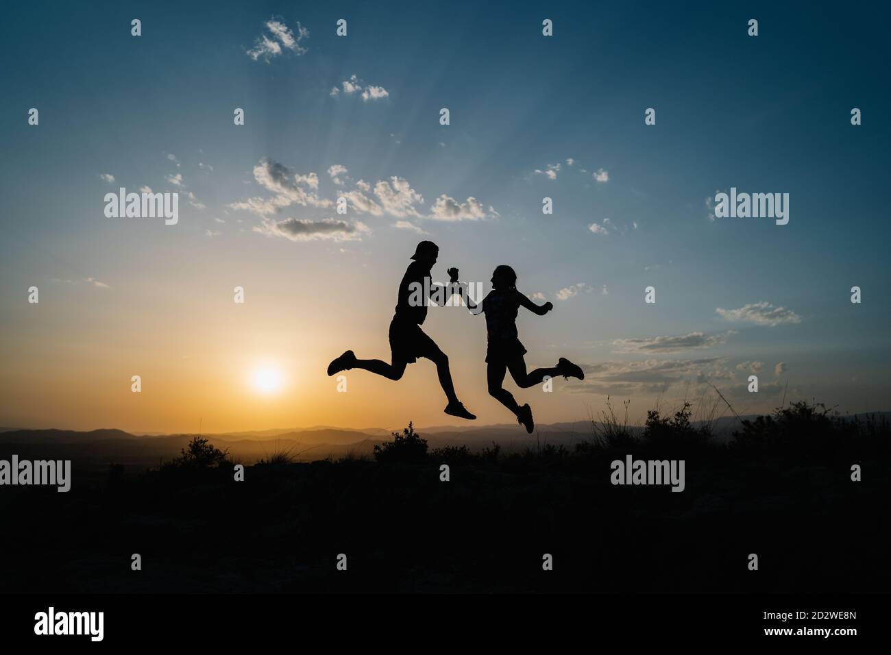 Side view silhouettes of unrecognizable male and female friends jumping high above ground while having fun and enjoying freedom in hilly terrain against sunset sky Stock Photo