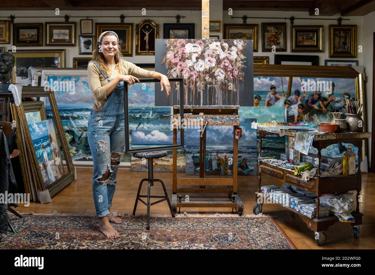 Full length of creative young female artist in denim overall holding empty frame while standing near easel with picture in workshop with paintings Stock Photo