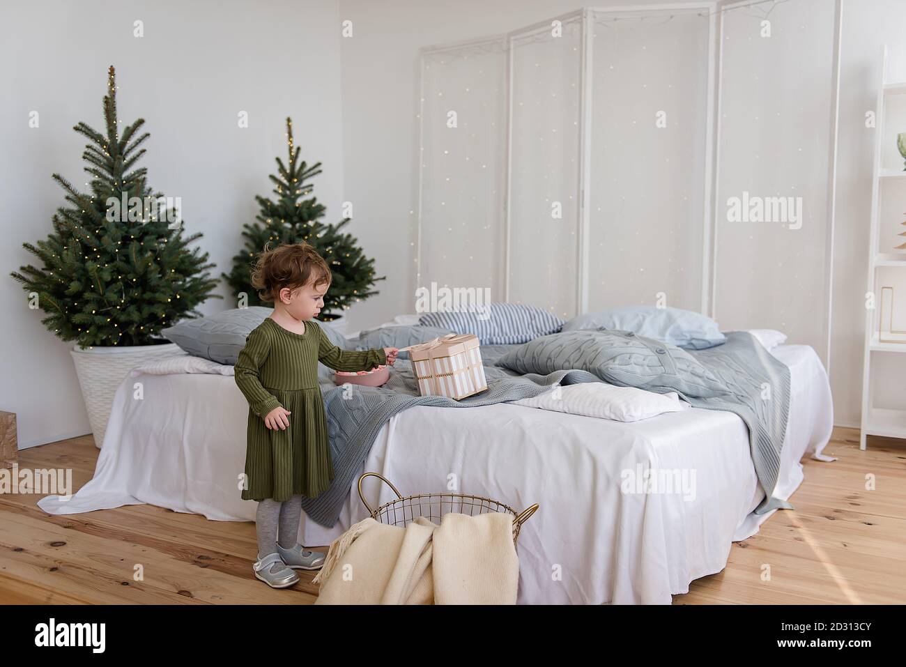 A little girl in a warm green dress found a present from Santa Claus on the sofa. Background in  blurred garland of lights, Christmas tree. White bed, Stock Photo