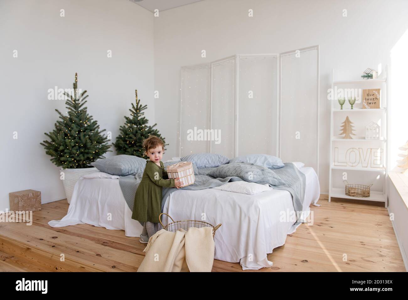 A little girl in a warm green dress found a present from Santa Claus on the sofa. Background in  blurred garland of lights, Christmas tree. White bed, Stock Photo