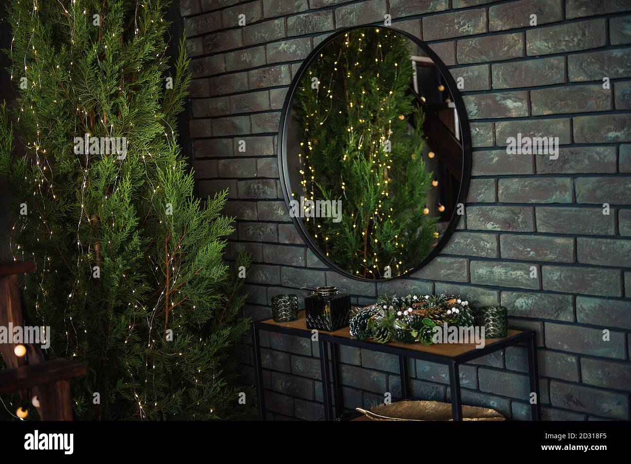 On a khaki brick green wall hangs a round mirror with a black frame. Around the Christmas trees in garlands with lights, juniper. Wallpaper Copy space Stock Photo